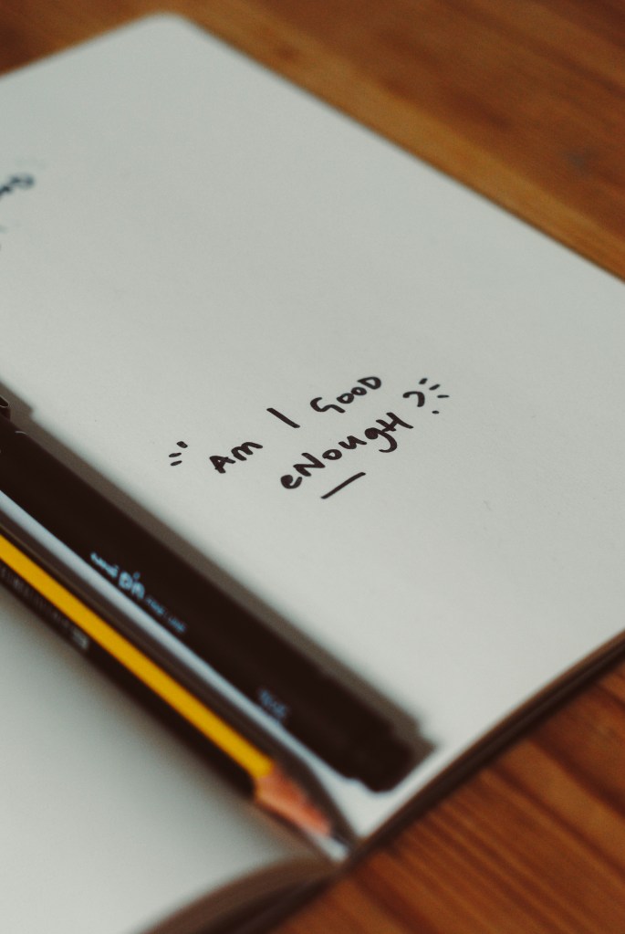 A notebook with handwritten text reading 'Am I good enough' in black ink on the page, with some lines for emphasis.