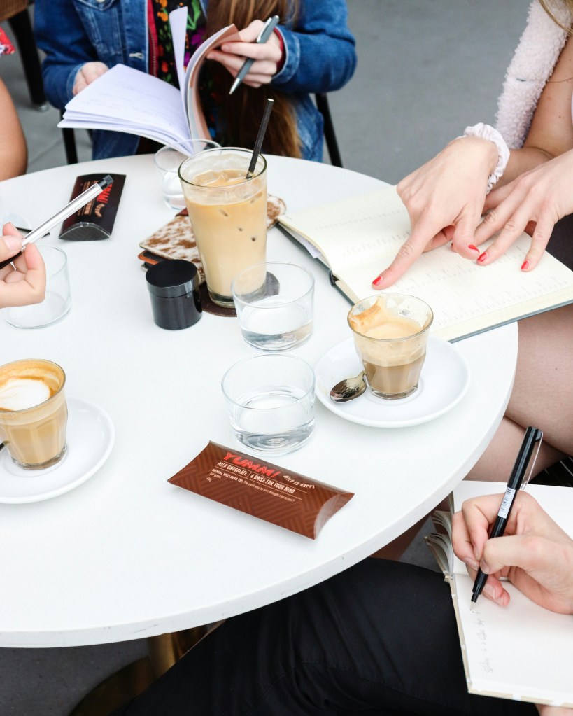 Image of a table with mulitple cups of coffee, and people writing in notebooks.
