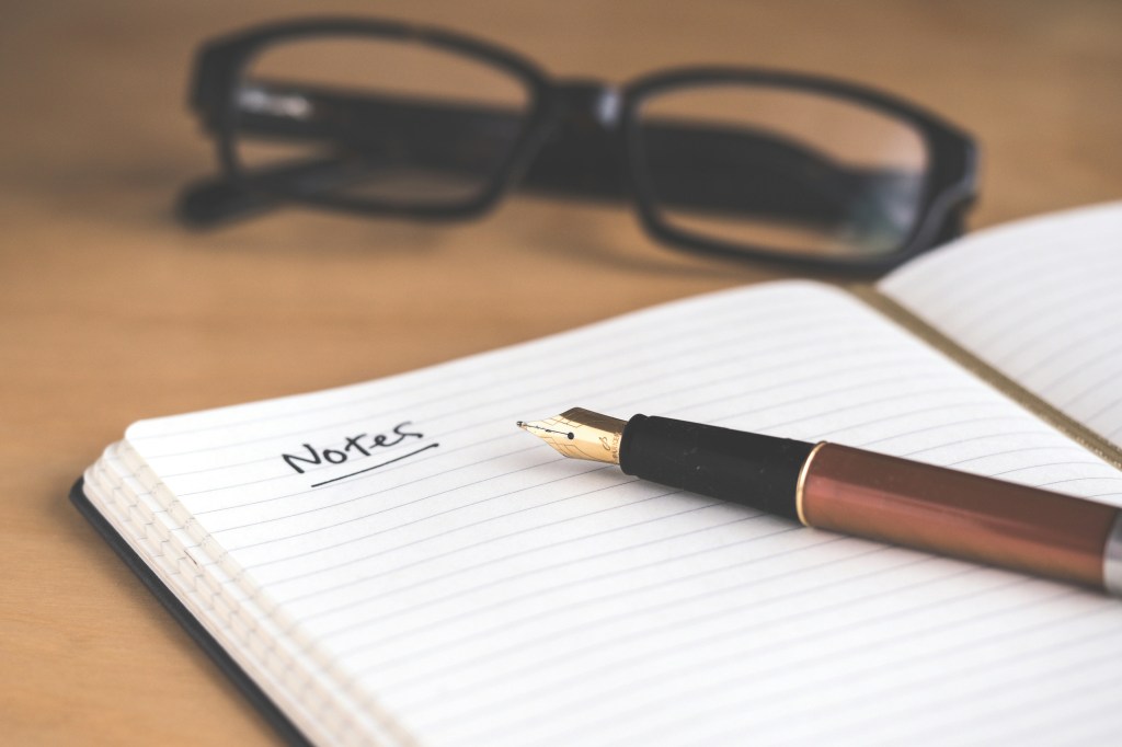 An image of a notebook open on a wooden desk. A fountain pen lays on a lined page. In the background there are black rimmed glasses that are out of focus. The page has 'notes' written and underlined, but the rest of the page is blank.