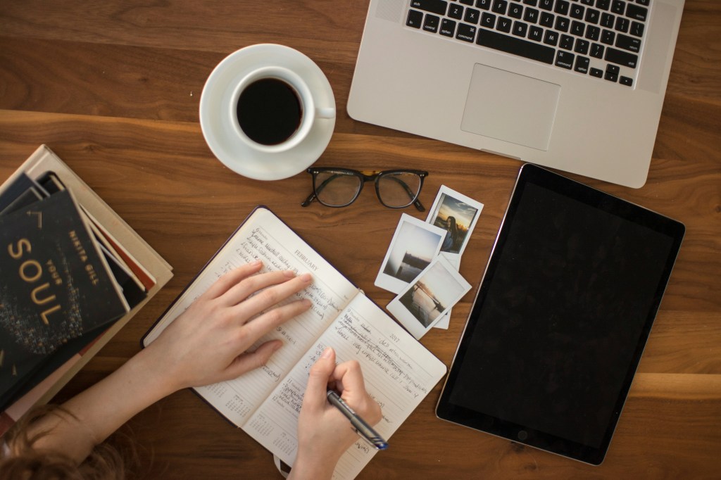 An overhead image of a cluttered desk as someone works. They are writing detailed notes in a notebook. They have multiple books to their left to reference. They have pictures, a laptop and a tablet all within easy reach, ready to find the information they need.